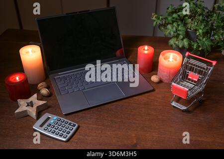 Laptop, calculator and a small shopping cart surrounded by candles and Christmas decoration on a dark wooden office desk, online shopping budget and h Stock Photo