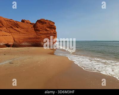 A peaceful beach bordered by a red sandstone cliff, under a clear blue sky. The waves gently caress the golden sand at the edge of the sea. Stock Photo