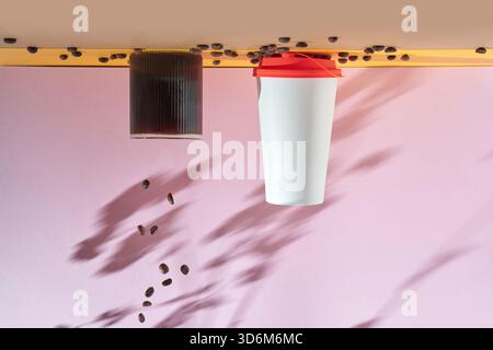 Creative coffee composition with a glass mug, reusable to go cup with orange lid and scattered roasted coffee beans casting soft shadows on a bright pink background Stock Photo