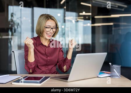 Excited businesswoman celebrating an online achievement, making a fist pump gesture while looking at her laptop screen, feeling joy and success at her corporate office desk Stock Photo