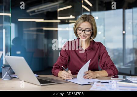 Businesswoman with glasses smiling while reviewing documents and notes, taking down details, and organizing information at her spacious, bright office workspace Stock Photo
