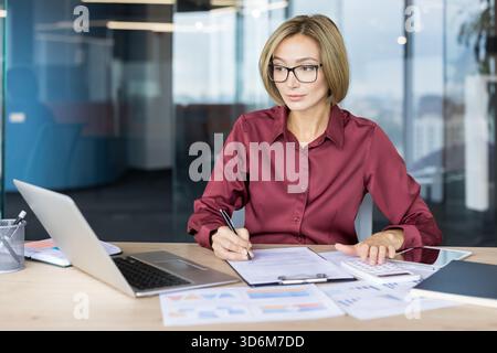 Professional businesswoman in glasses calculating expenses and writing notes on documents, managing finance and accounting tasks at a modern office desk with laptop and charts Stock Photo