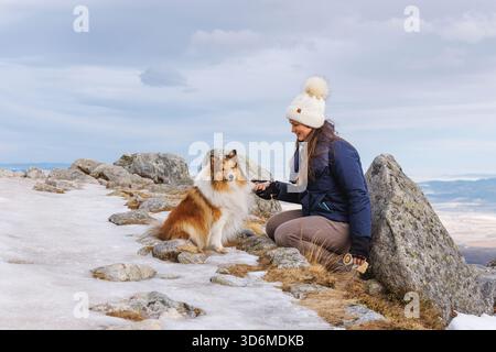 A woman kneels beside her collie on a snowy mountaintop. Rocks and frost form a rugged high-altitude landscape under soft light. Stock Photo