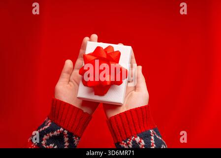 Top view of hands holding a white gift box with a red bow on a bright red background, perfect for festive campaigns and holiday creatives Stock Photo