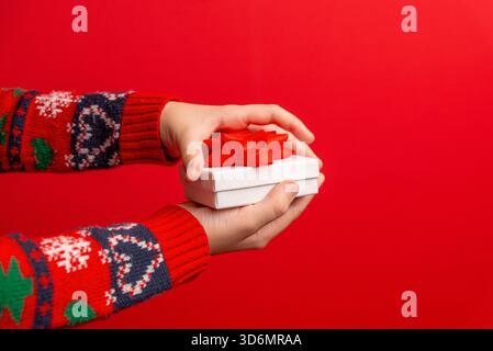 Hands holding a small white gift box with a red bow on a vivid red background, ideal for festive branding, holiday sales, and promotional graphics Stock Photo