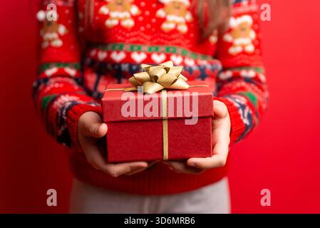 Child holding a red gift box with a gold bow against a bright red background, perfect for holiday themes, festive marketing, and seasonal visuals Stock Photo