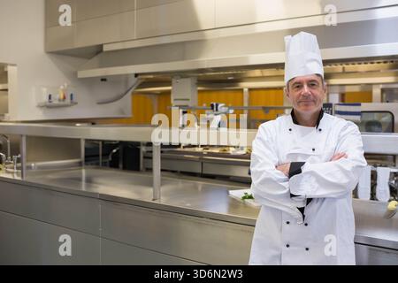 Middle-aged chef in chef-coat, tall hat standing arms crossed by steel pass in kitchen, copy space. Stainless, professional, galley, service, prep, ho Stock Photo