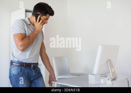 Adult male standing holding phone, leaning on white desk with screen in home office, copy space. Minimalist, modern, bright, workspace, workstation, m Stock Photo