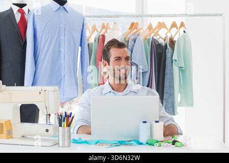 Man working at white table in fashion studio, using laptop and sewing machine near mannequins. Designer, atelier, workspace, garments, fabrics, thread Stock Photo
