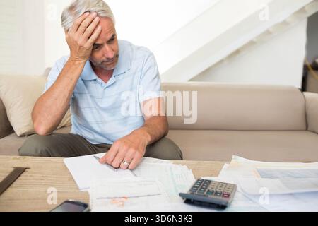 Senior man sitting on beige sofa wearing blue polo, reviewing bills on wooden table with calculator. Envelopes, receipts, papers, phone, focus, daylig Stock Photo