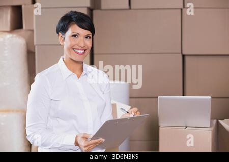 Mature woman standing in storage area wearing white shirt holding clipboard and pen near laptop. Warehouse, logistics, inventory, supervisor, fulfillm Stock Photo