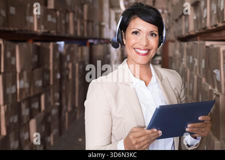 Mature female manager standing in narrow warehouse aisle holding dark tablet and using headset. Storage, facility, logistics, inventory, shelving, box Stock Photo