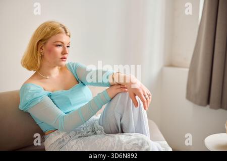 Young woman in light blue top sitting on sofa, looking pensive Stock Photo