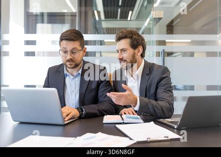 Two businessmen discussing project details and analyzing data on a laptop, fostering teamwork and professional collaboration in a modern office environment Stock Photo