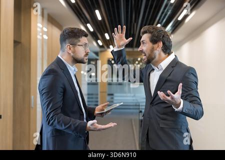 Two male colleagues arguing intensely in a modern open-plan office, shouting and gesturing in a heated professional dispute while one holds a tablet, showing stress and tension Stock Photo