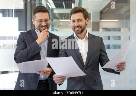 Two bearded businessmen in suits smiling and reviewing financial reports together in a modern office, collaborating on strategy, planning and paperwork during a meeting Stock Photo