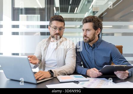 Two male business partners discussing work and reviewing data on a laptop, actively brainstorming ideas and planning a new project during an office meeting Stock Photo