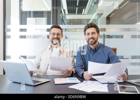 Two smiling colleagues review documents and a laptop at a modern office desk, collaborating on reports and strategy during a productive, friendly business meeting Stock Photo