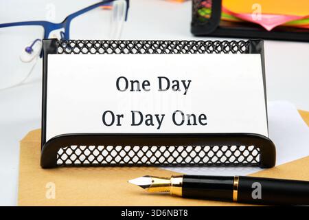 The desk shows a framed message card that reads One Day Or Day One, set near glasses and a pen, suggesting a motivational work environment or a moment Stock Photo