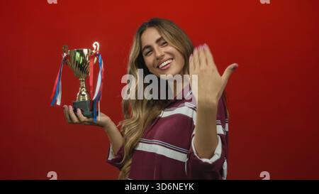 Woman holds trophy in studio with finger pointing to trophy highlighting the cup against red background; achievement celebration success pride. Stock Photo