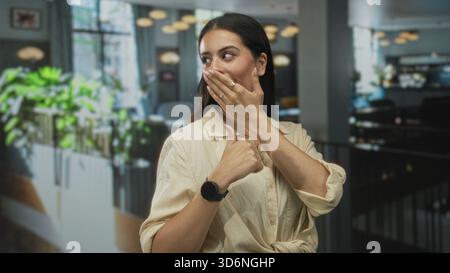 Young brunette woman covering mouth with hand and thumb pointing to herself in restaurant building; surprise playful moment. Stock Photo