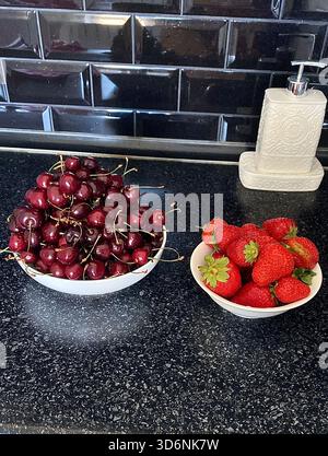 Two bowls of ripe strawberries and cherry close to the white soap dispenser at the black kitchen Stock Photo