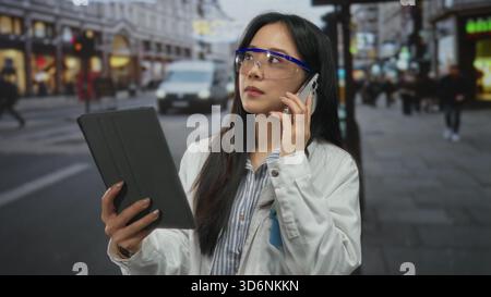 Woman scientist using tablet and phone outdoors on busy urban street, wearing safety goggles and white coat, exemplifying multitasking and technology Stock Photo