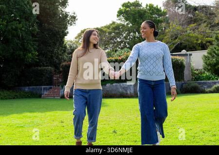 Diverse women walking hand in hand across garden lawn beside stone wall shrubs and brick steps Stock Photo