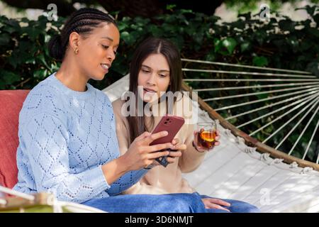 Mother and daughter sitting on hammock in yard checking credit card on smartphone drinking iced tea Stock Photo