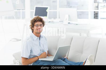 Adult male sitting on light gray sofa in office lounge using laptop, wearing glasses and shirt. Coworking, daylight, minimalist, modern, monitor, whit Stock Photo