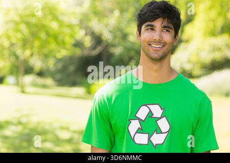 Adult Asian male standing and smiling in park, wearing green tee with recycling logo, copy space. Person, outdoors, volunteer, nature, canopy, sunlit, Stock Photo