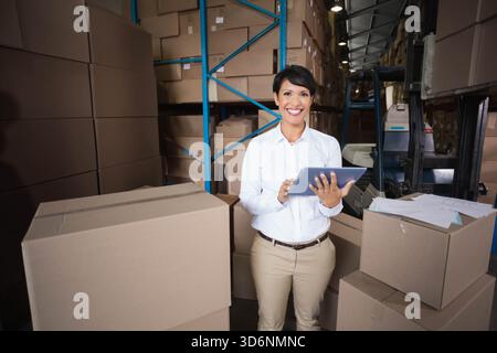 Female warehouse supervisor standing and tapping tablet in aisle with boxes papers in white shirt. Woman, manager, logistics, storage, inventory, faci Stock Photo