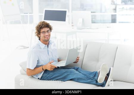 Man reclining on white leather sofa in bright lounge using silver laptop, wearing light-blue shirt. Gentleman, office, modern, minimalist, workspace, Stock Photo