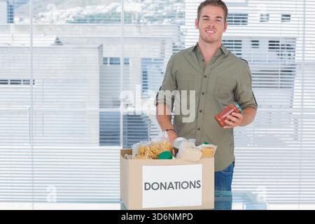 Male standing in green shirt holding jar over DONATIONS box at office, copy space. Volunteer, groceries, nonperishable, urban, daylight, community, ou Stock Photo