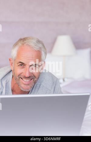 Mature male leaning forward and typing on laptop in bedroom, wearing light gray top. Senior, man, device, keyboard, silver, bed, headboard Stock Photo