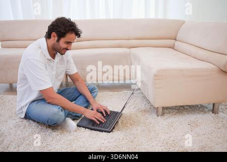 Mid-adult man sitting cross-legged on ivory shag rug typing on black laptop in living room. Remote, work, home, office, computer, daylight, minimalist Stock Photo