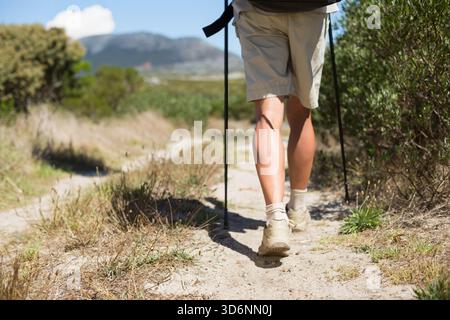 Male hiker walking on narrow dirt trail using poles wearing shorts hiking shoes toward mountains. Path, arid, scrubland, backpack, sunlit, shadow, hor Stock Photo