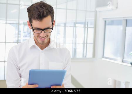 Man in white shirt black glasses viewing blue tablet in bright office with grid windows. Minimalist, highkey, professional, workspace, device, screen, Stock Photo