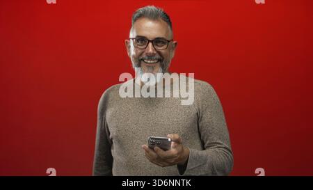 Man holding smartphone and smiling, right hand visible, wearing glasses with grey beard and brown sweater in a red studio; relaxed connection. Stock Photo