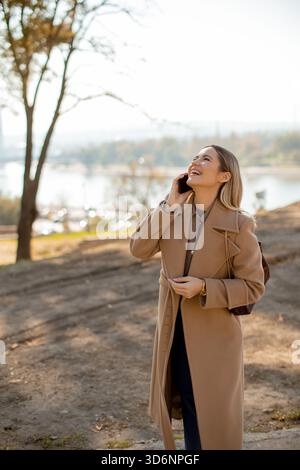 A woman in a light brown coat engages in a joyful phone conversation. She stands on a grassy slope, surrounded by trees, with a river visible in the b Stock Photo