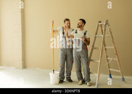 Two men in work overalls are preparing to paint a room with a light brown wall. One holds a paint roller while the other looks at a clipboard, suggest Stock Photo
