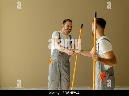 Two men in gray work overalls smile and shake hands in a clean, freshly renovated room. They are holding long paint rollers, ready to start their pain Stock Photo