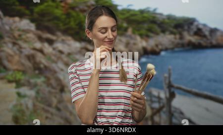 Woman holding paintbrushes and touching chin at seaside near rocky coast in striped shirt and braid; contemplation. Stock Photo
