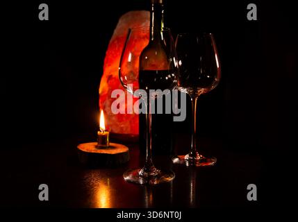 Two elegant empty clear wineglasses for red wine with wine bottle on the table with burning candle on dark black background with blur orange salt lamp Stock Photo