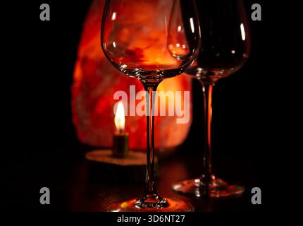 Two elegant empty clear wineglasses for red wine on the table with burning candle on dark black background with blur orange salt lamp. Closeup decor c Stock Photo