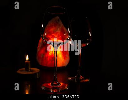 Two elegant empty clear wineglasses for red wine on the table with burning candle on dark black background with blur orange salt lamp. Closeup decor c Stock Photo