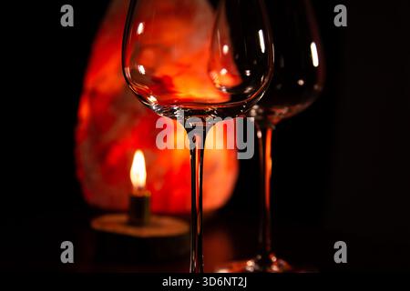 Closeup two elegant empty clear wineglasses for red wine on the table with burning candle on dark black background with blur orange salt lamp. Still l Stock Photo