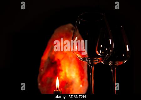Closeup two elegant empty clear wineglasses for red wine on the table with burning candle on dark black background with blur orange salt lamp. Still l Stock Photo