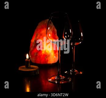 Two elegant empty clear wineglasses for red wine on the table with burning candle on dark black background with blur orange salt lamp. Closeup decor c Stock Photo