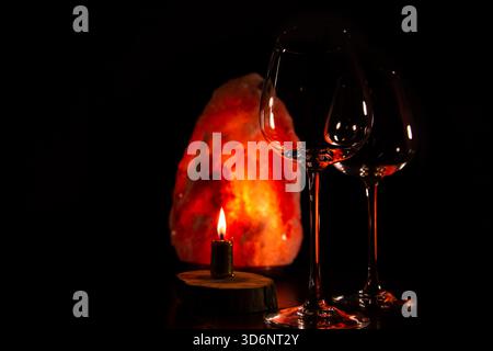 Two elegant empty clear wineglasses for red wine on the table with burning candle on dark black background with blur orange salt lamp. Closeup decor c Stock Photo